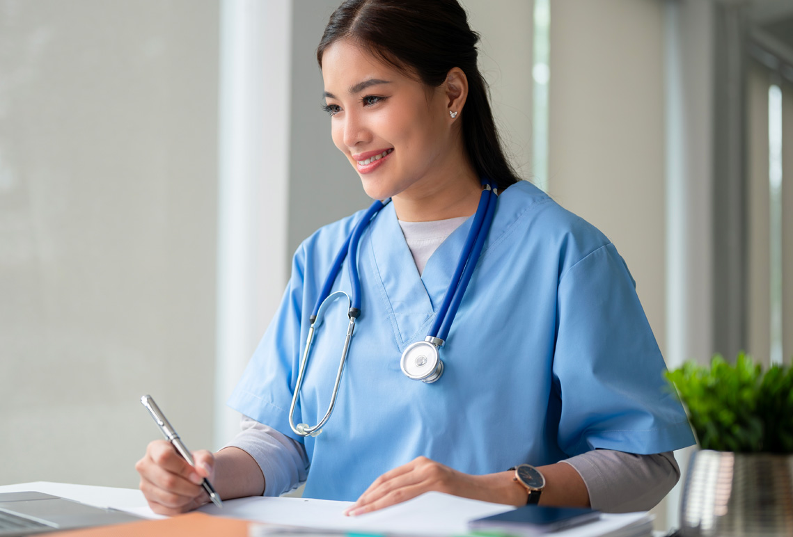 A student in blue scrubs and with stethoscope makes notes while sitting at a desk.